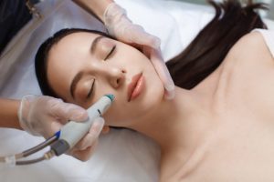 A woman with long brown hair lies on her back with her eyes closed, receiving a relaxing facial treatment using a skincare device on her cheek. The hands of the professional performing the treatment are gloved. Experience top-notch skin care in Wake County for radiant results.