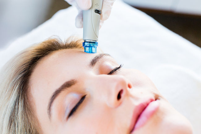 A person is lying down with eyes closed, receiving a Hydra Facial treatment. A gloved hand is holding a device with a blue light near the person's forehead, suggesting an advanced skincare procedure. The overall setting appears to be a clinical or spa environment in Wake County.
