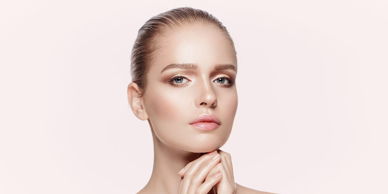 A woman with fair skin and blue eyes gazes into the camera, her neutral expression complemented by slicked-back hair, suggesting she might have just enjoyed a refreshing Hydra Facial in Wake County. She rests her chin lightly on her hands, with soft makeup emphasizing her eyes and lips. The background is light and plain.