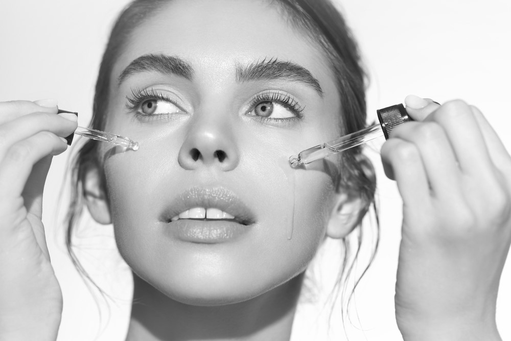 Black and white photo of a person with clear skin, applying a cosmetic serum to their face with two dropper applicators. The person gazes slightly upward, with a serene expression, embodying the essence of Skin Care Wake County. The background is plain and light.