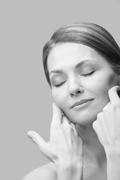A grayscale image of a woman with closed eyes, gently touching her face with both hands. She appears relaxed and peaceful, showcasing the smooth skin often achieved through Hydra Facials Wake County. Her hair is pulled back and the background is a plain light gray.