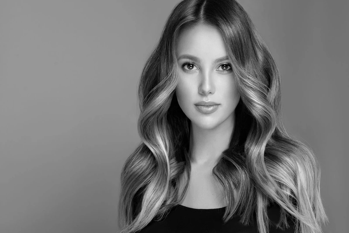 Black and white portrait of a young woman with wavy hair cascading past her shoulders. She has a calm, confident expression, wearing a simple, dark top. The background is plain and light gray, emphasizing her soft features and natural beauty—reflecting the excellence of skin care in Wake County.