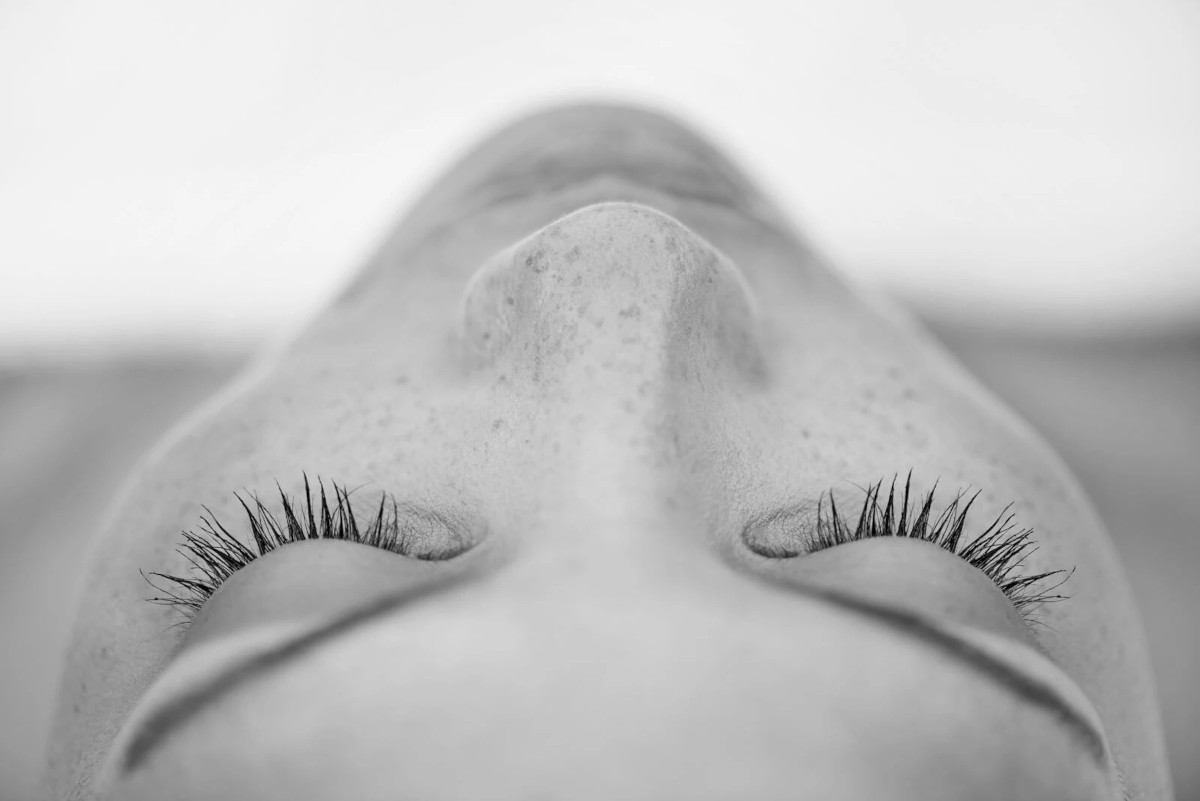 Black and white close-up photo of a person's face taken from above. The person has their eyes closed, displaying long eyelashes. Their skin, possibly touched by Microdermabrasion Wake County treatments, has a few freckles. The shot captures the subtle contours of the nose and forehead. The background is blurred.