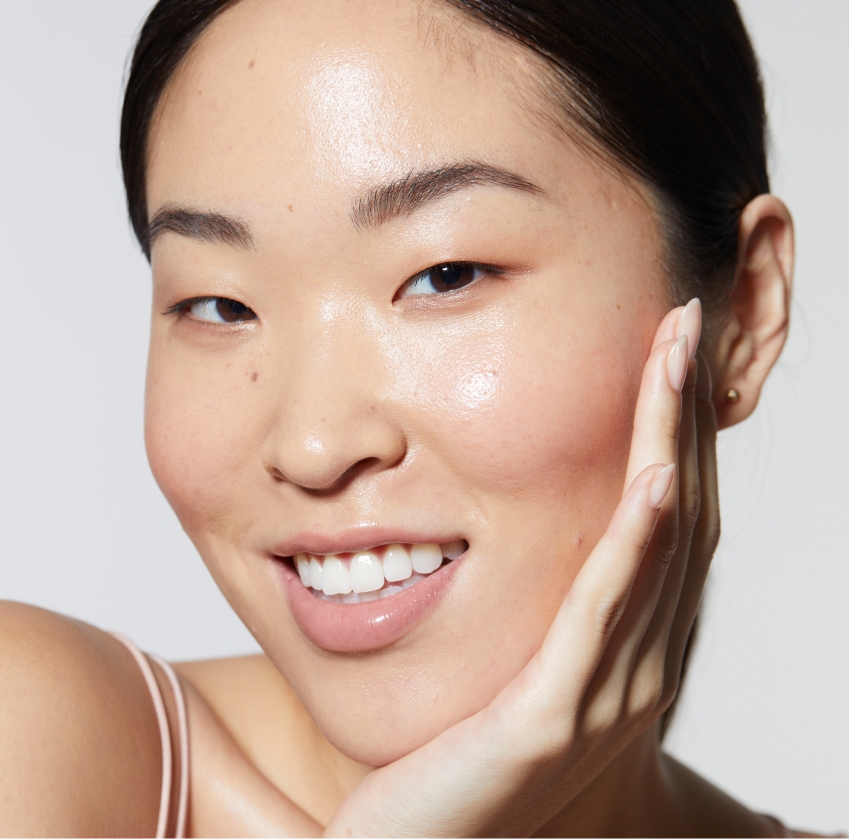 A person with clear, glowing skin from Hydra Facials Wake County, dark hair pulled back, and minimal makeup smiles with one hand gently touching their cheek. They are wearing a light pink top against a plain background.
