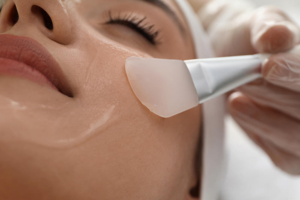 Close-up of a person's face receiving a Hydra Facial treatment. A smooth, rounded applicator held by a gloved hand is applying a clear, gel-like substance on their cheek. The person has their eyes closed and is resting comfortably.