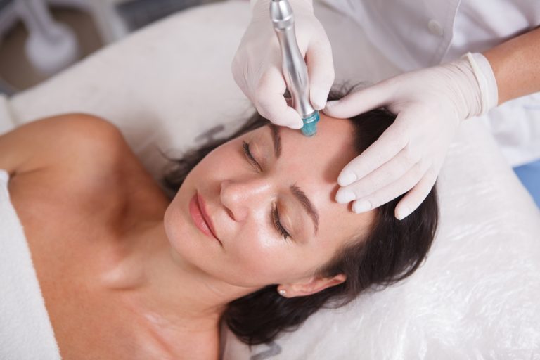 A woman is lying on a bed with her eyes closed, receiving a Hydra Facials Wake County treatment. A professional wearing white gloves is using a handheld device on her forehead. The woman looks relaxed, with a slight smile on her face.