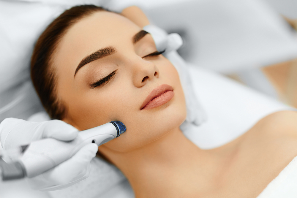 A woman with closed eyes is receiving a hydra facial treatment on her face. She appears relaxed while a professional in white gloves uses an electronic device on her cheek. The scene is set in a clinical or spa-like environment in Wake County.