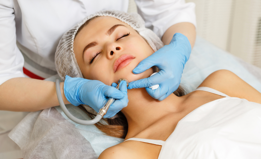 A person is receiving a Hydra Facial treatment from a professional in Wake County. They are lying down with their eyes closed as the expert, wearing blue gloves, uses a device on their face. The person has a white head cover and is covered with a white towel.