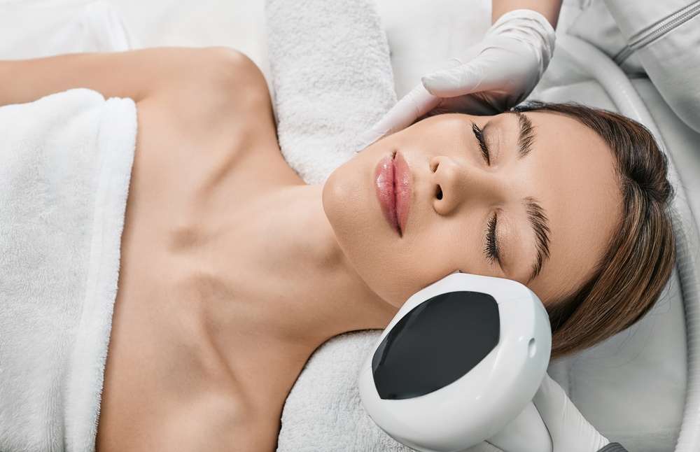 A woman lies on a massage bed with her eyes closed, receiving a beauty treatment. A gloved hand holds a white handheld device to her face for microdermabrasion Wake County. She appears relaxed, wrapped in a white towel, with a serene expression. Medical devices and towels are visible.