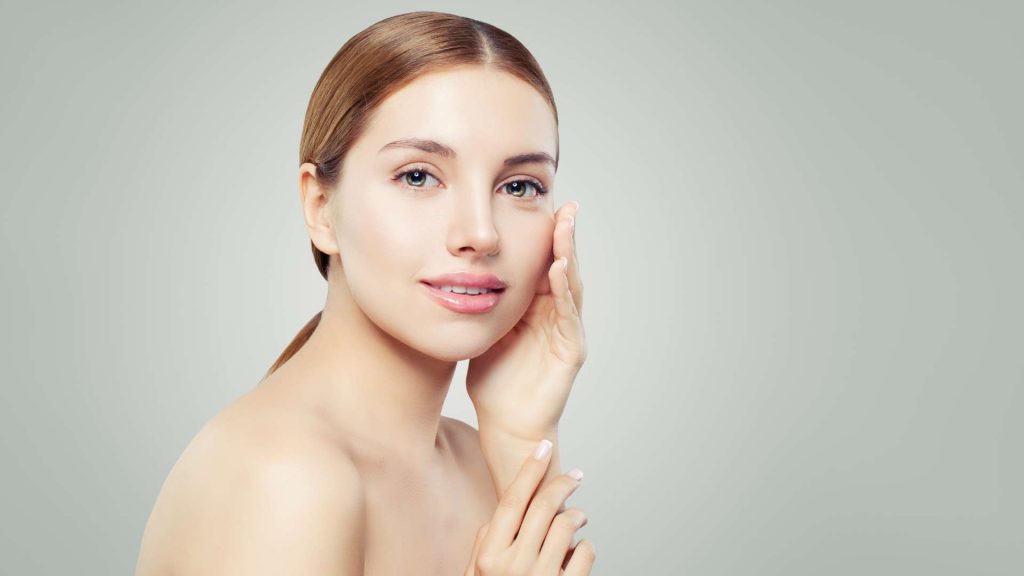 A woman with fair skin and light brown hair is posing against a plain background. She is looking into the camera with a serene expression, one hand touching her cheek and the other raised near her chin. With bare shoulders and natural makeup, she showcases the smooth results of Hydra Facials Wake County.
