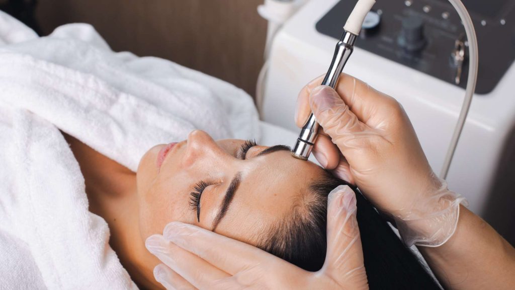 A person undergoing a facial treatment. A practitioner wearing gloves uses a handheld device on the person's forehead, while the person lies down with eyes closed, covered in a white towel. The background shows medical equipment, highlighting advanced skin care in Wake County.