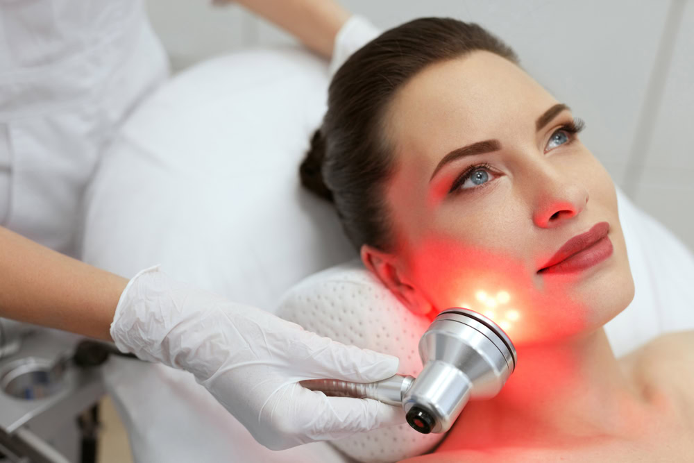 A woman receives a Hydra Facial in a clinic in Wake County. She lies on a cushioned bed while a gloved professional expertly holds a device emitting red light against her cheek. The woman looks relaxed and content, surrounded by the clean, clinical setting focused on top-tier skincare.