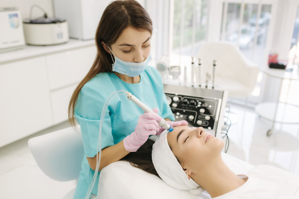 A beautician wearing a surgical mask and pink gloves performs a Hydra Facial on a reclining woman with a white headband in a modern, well-lit clinic. Medical equipment and a large window are visible in the background, showcasing the professional skin care available in Wake County.