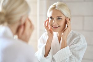 A woman with blonde hair smiles as she looks at herself in a bathroom mirror. She is wearing a white bathrobe and gently touching her face with both hands, appearing relaxed and content, likely after one of the soothing Hydra Facials Wake County offers. The background shows tiled walls, giving a clean and serene atmosphere.