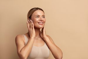 A woman with her hair pulled back is smiling and gently touching her face with both hands. She is wearing a beige tank top, and the background is a solid light brown color. Her expression is calm and content, as if she just enjoyed one of the luxurious Hydra Facials in Wake County.