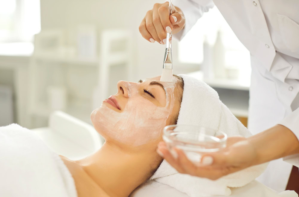 A person lies on a spa bed with a white towel wrapped around their head. Another individual is applying a white facial mask to their face using a brush. The setting is bright and serene, suggesting a relaxing Skin Care Wake County environment.
