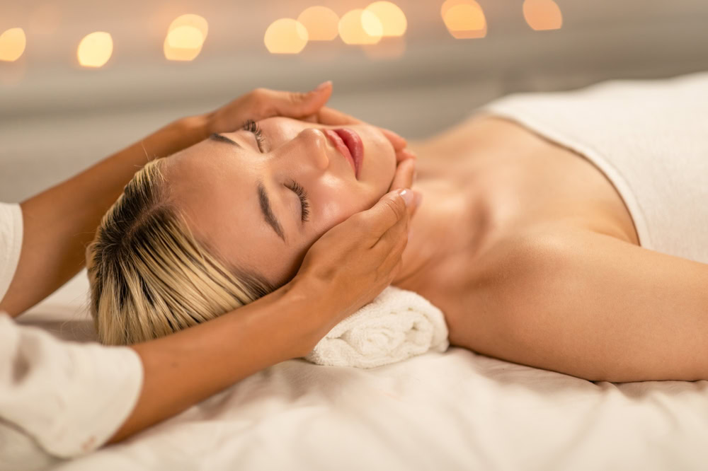 A person with closed eyes lies on a white towel while receiving a facial massage. Soft, warm lighting in the background creates a serene and relaxing atmosphere, perfect for Hydra Facials Wake County. The person appears calm and comfortable.
