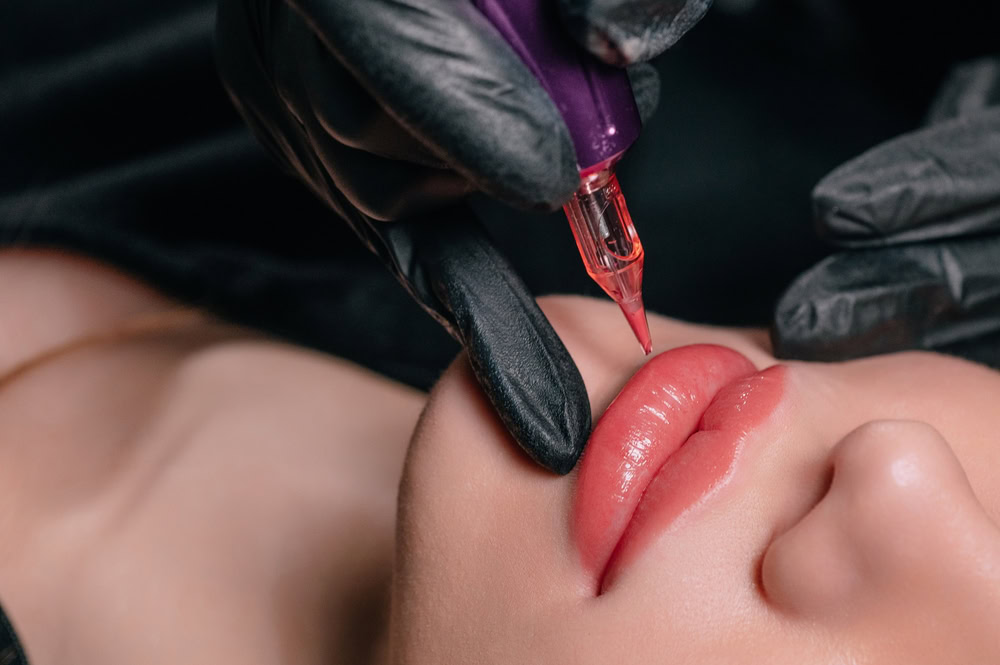 A close-up of a person's lips being tattooed by a professional wearing black gloves. A tattoo machine with red pigment is adding color to the lips, creating a cosmetic lip tattoo. The person receiving the tattoo has light skin and slightly parted lips, perfect for their upcoming Hydra Facials Wake County session.
