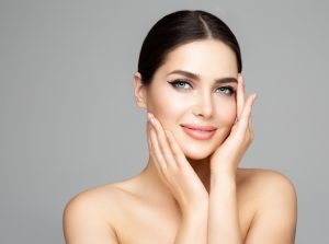 A woman with dark hair, slicked back in a bun, smiles gently with her hands touching her cheeks. Her clear skin and light makeup reflect the impeccable results of premium skin care in Wake County. She wears no visible clothing against a plain background.
