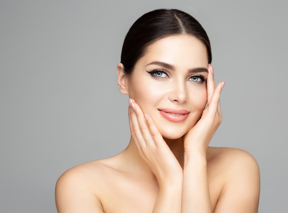 A woman with dark hair, slicked back in a bun, smiles gently with her hands touching her cheeks. Her clear skin and light makeup reflect the impeccable results of premium skin care in Wake County. She wears no visible clothing against a plain background.