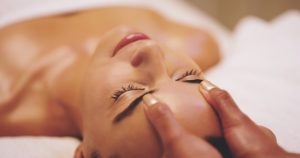 A person is lying down with eyes closed, receiving a facial massage. Hands are gently pressing on their forehead. The setting appears calm and relaxing, suggesting a spa or wellness environment specializing in Skin Care in Wake County.