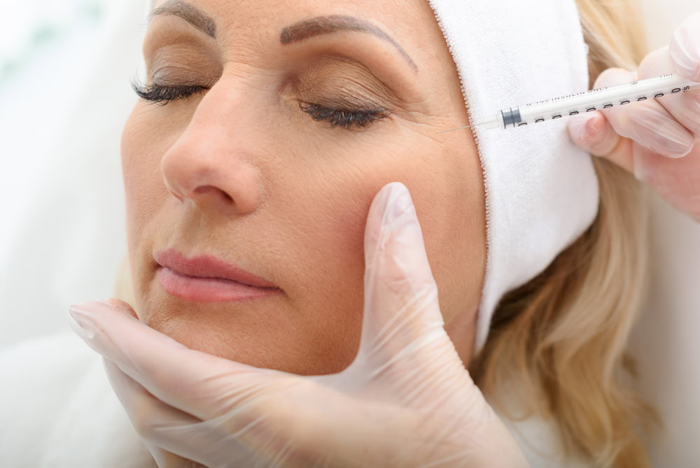 A close-up of a person with a serene facial expression, eyes closed, receiving a cosmetic injection near the outside corner of their eye. The individual administering the injection wears gloves and holds the patient's face gently with one hand, offering exceptional skin care in Wake County.