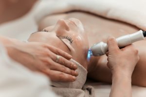 A person receives a facial treatment while lying down, with their hair covered by a towel. A healthcare professional gently holds the person's chin for support while using a handheld device emitting blue light. This session is part of the comprehensive Hydra Facials Wake County offers for rejuvenated skin.