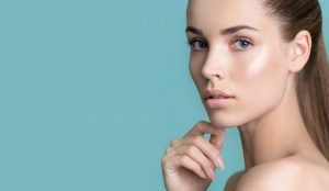 A close-up of a person with fair skin, blue eyes, and long, light brown hair pulled back, posing against a solid light blue background. Their face is slightly turned to the side, and they are gazing directly at the camera with a thoughtful expression. Achieve such flawless skin through expert Skin Care Wake County services.