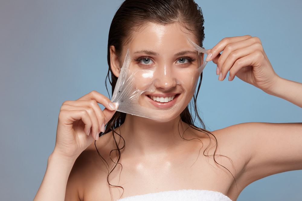 A woman with wet hair smiling while peeling off a clear face mask. She is wrapped in a white towel against a light blue background.