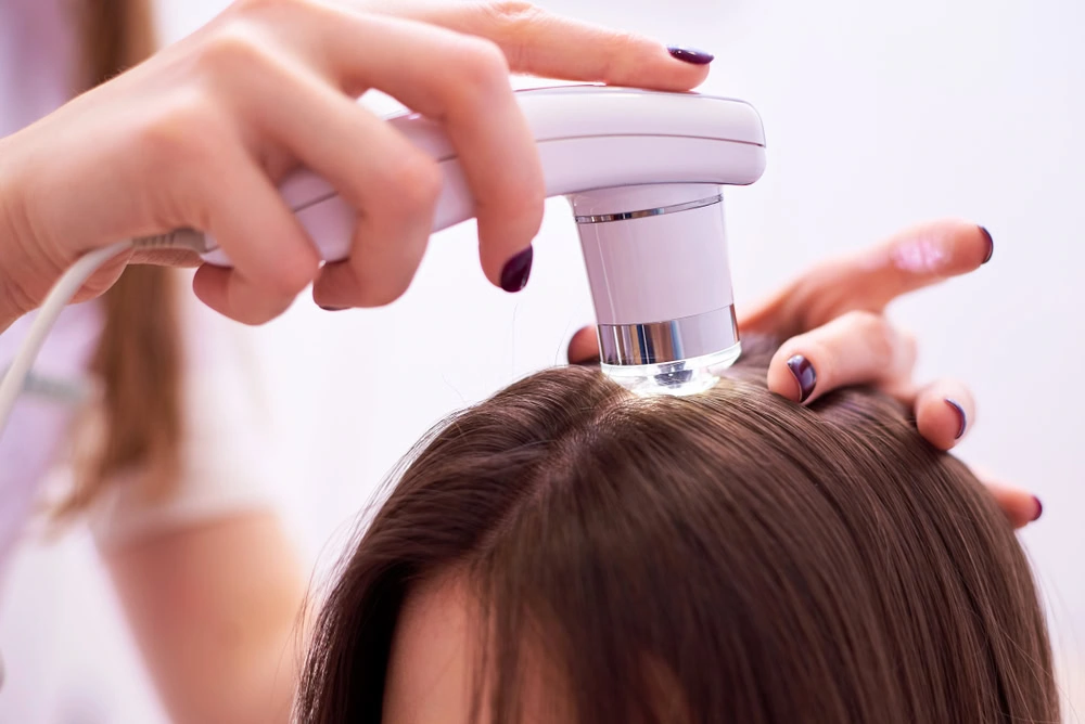 A person is receiving a scalp treatment with a handheld device. The technician's hand holds the device close to the subject's hair, focusing on the roots. The setting is a clinical or salon environment with a neutral background.