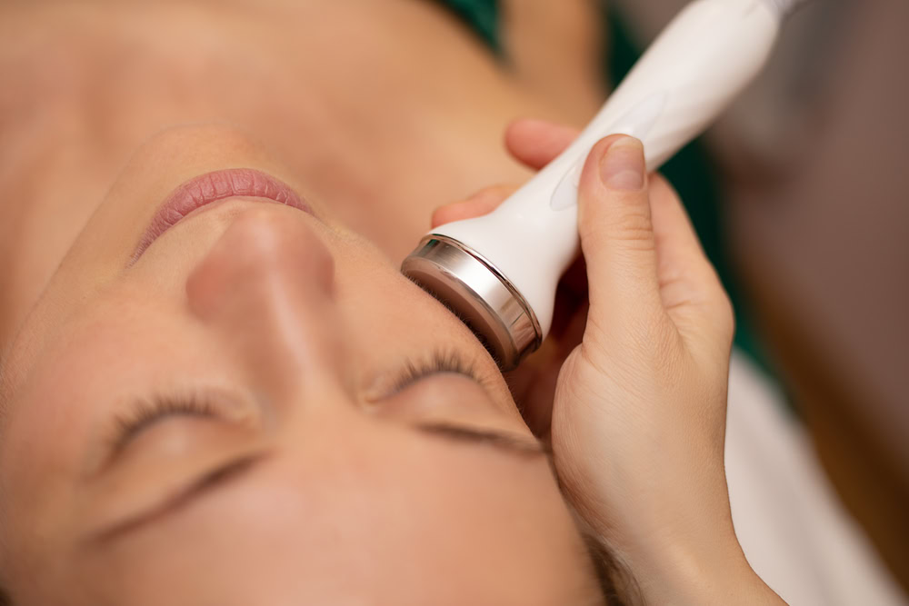 A person receiving a facial treatment with a spa device. The close-up shows the device being applied to the cheek, held by a hand. The person has closed eyes, suggesting a relaxing or soothing experience.