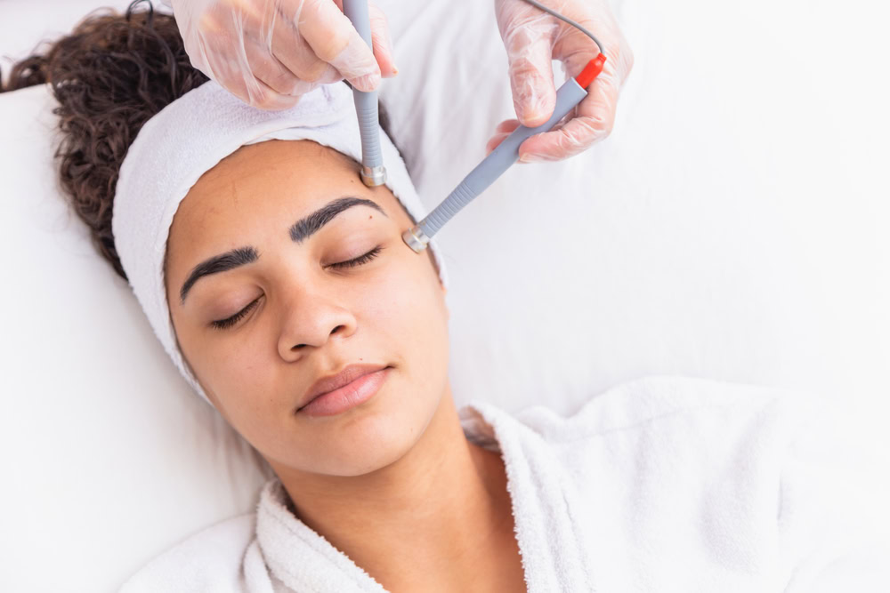 A person is receiving a microcurrent facial treatment. They lie relaxed on a treatment bed, eyes closed, with a white headband and a robe. Gloved hands hold devices near the person's temples. The setting appears calm and clinical.