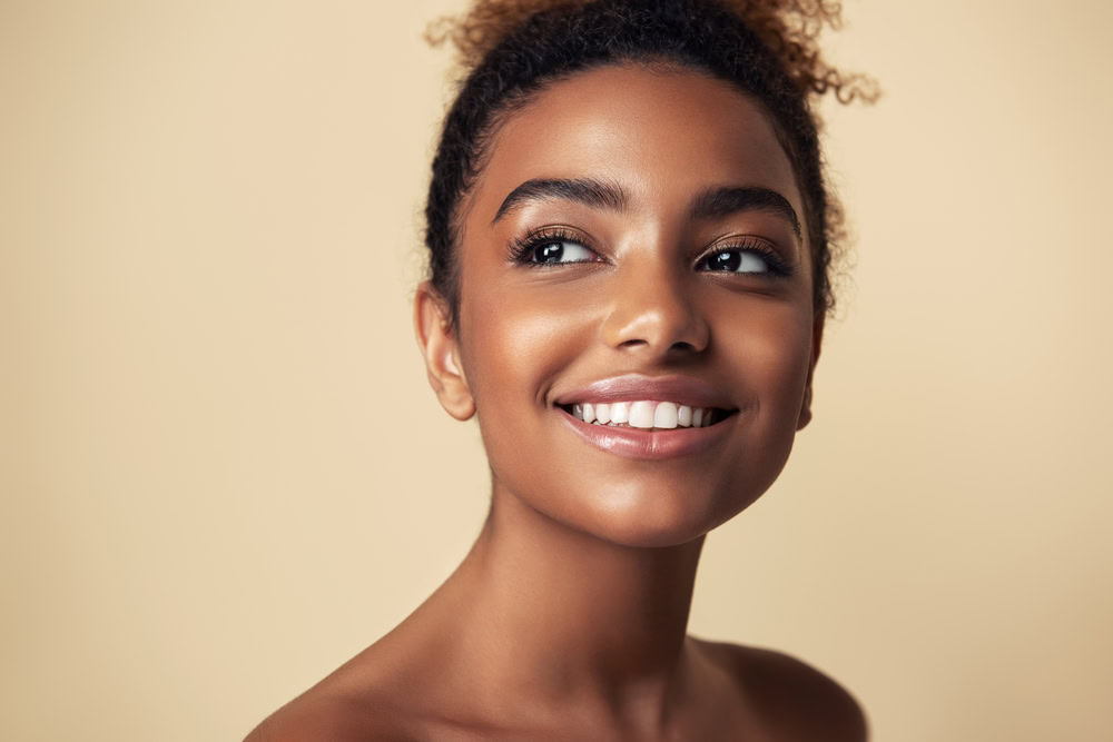 A person with curly hair smiles brightly against a beige background. Their skin glows, and they appear happy and relaxed.
