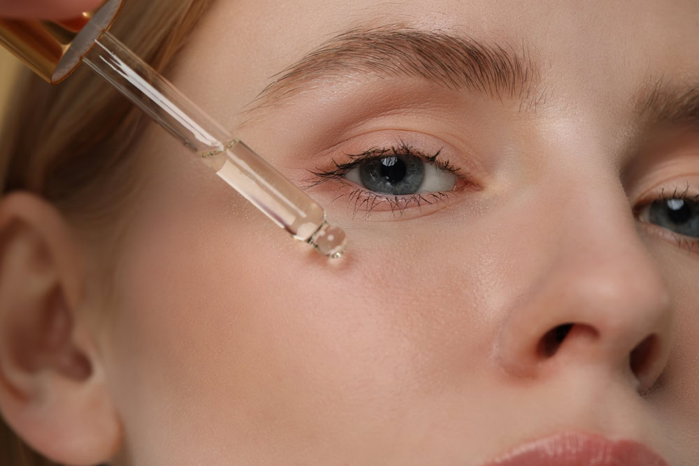 Close-up of a person applying facial serum with a dropper beneath their eye. The individual's skin is smooth, and their eyes are blue. The background is neutral, emphasizing the skincare routine.