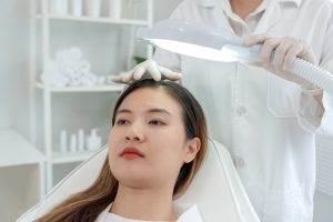 A woman sits in a beauty clinic, undergoing a scalp examination. A professional in a white coat and gloves holds a magnifying lamp above her head. Shelves with skincare products and towels are in the background.