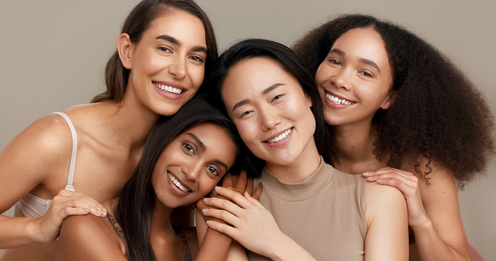 Four women smiling and standing close together, each with different hair and skin tones, against a neutral background. They appear joyful and relaxed, with their arms around one another.