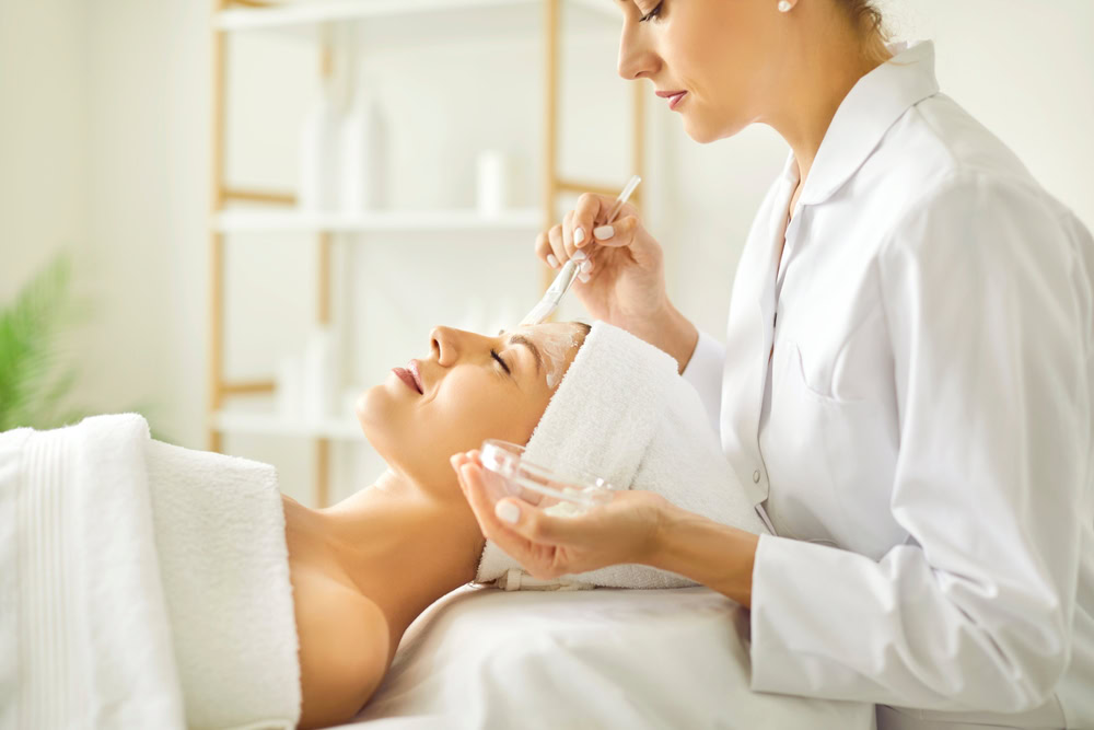 A person is lying down in a spa, covered in a towel, with a towel wrapped around their head. An esthetician in a white uniform is applying a facial treatment with a brush. The background features shelves with bottles.