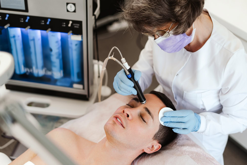 A person receiving a facial treatment at a spa or clinic. A professional in a white coat and gloves uses a handheld device on the client's forehead, who is lying down with eyes closed. A machine and products are visible in the background.