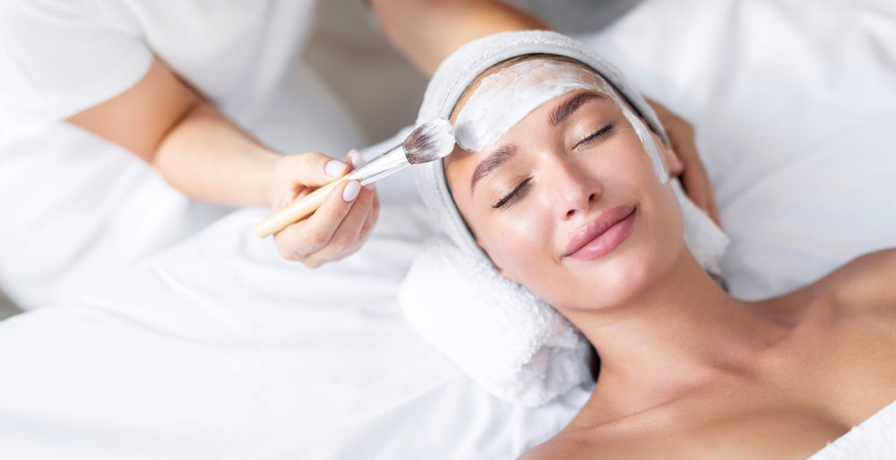 A person is lying down with a towel around their head, receiving a facial treatment. A hand applies a creamy mask to their forehead with a brush. The person appears relaxed and is smiling slightly, eyes closed.