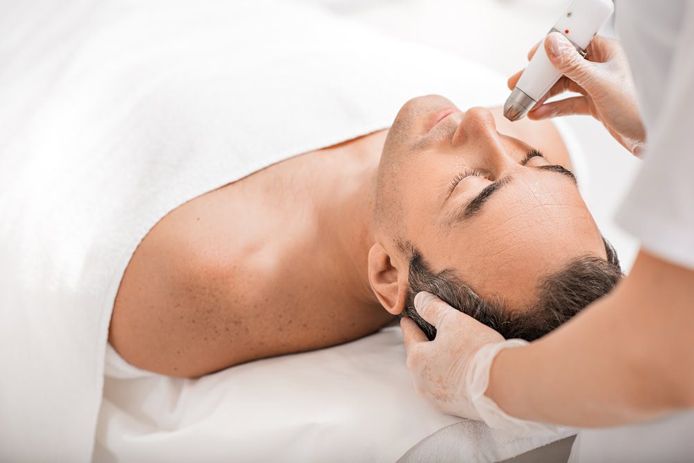 A person receives a facial treatment while lying on a spa bed, covered with a white towel. A gloved practitioner applies a hand-held device to the person's forehead in a serene setting.