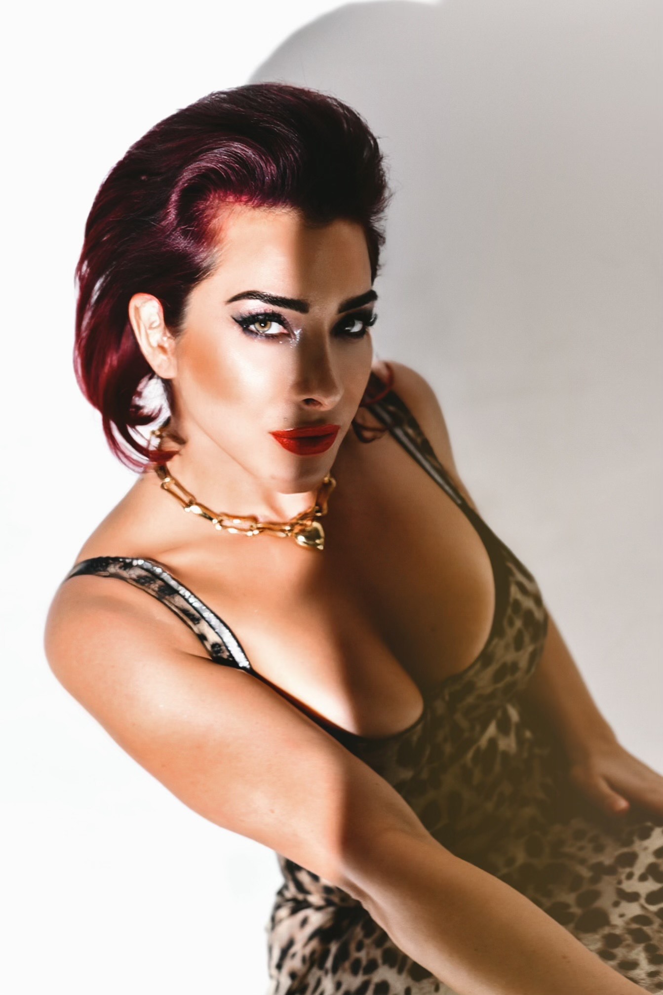 A striking woman with red hair, clad in a leopard print dress and gold necklace, exudes confidence against a white background. With bold makeup featuring red lipstick and winged eyeliner, she gazes intensely upward—a true embodiment of the empowerment championed by Wake Skincare.