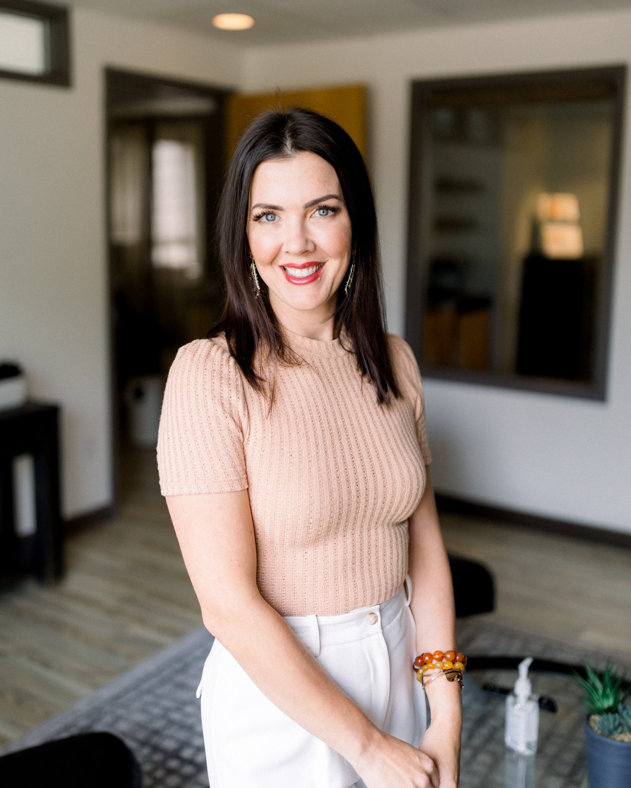 A woman with long dark hair wearing a beige short-sleeve top and white pants stands smiling in a modern office space with light wood floors and neutral decor, reflecting the fresh aesthetic of About Wake Skincare.