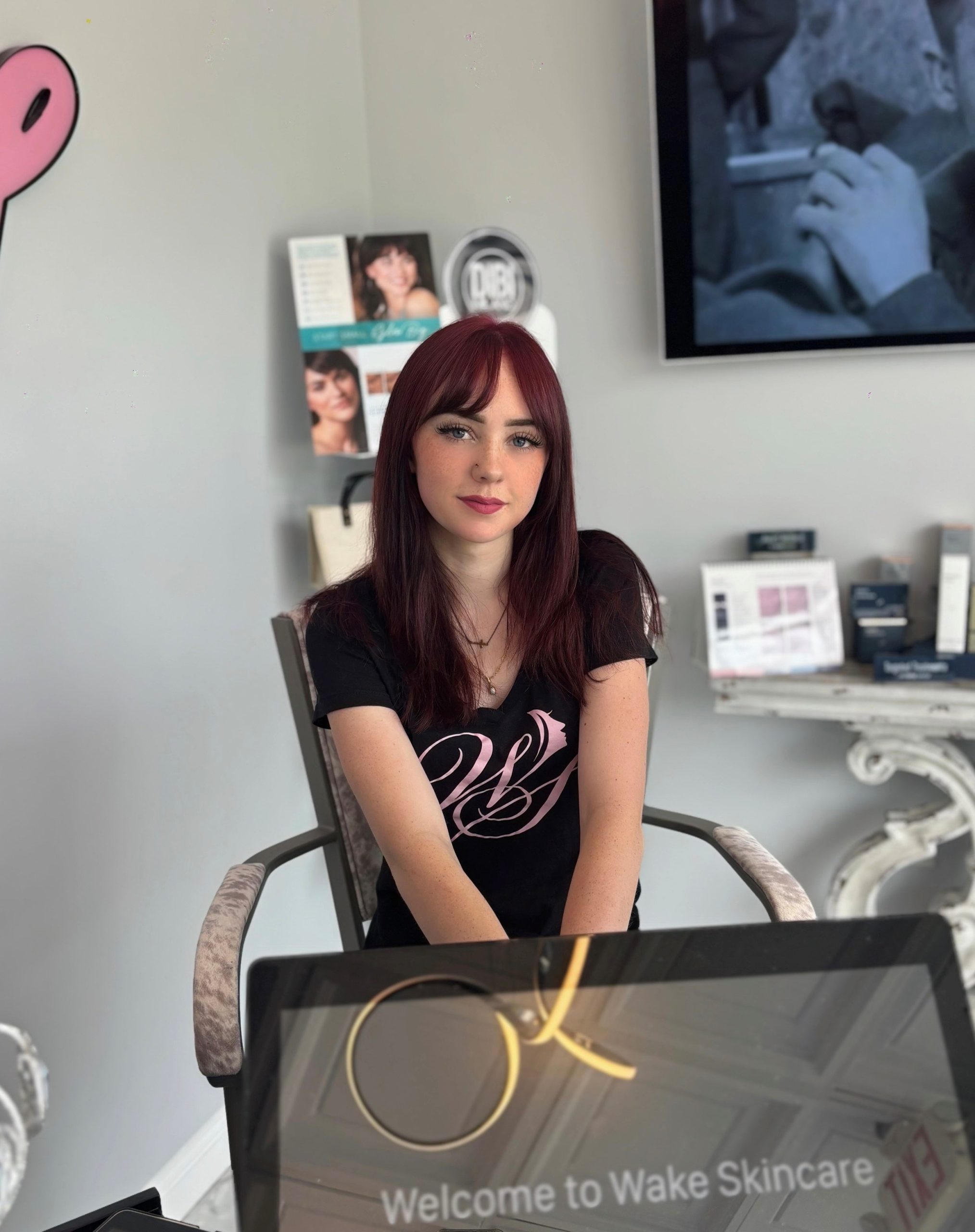 A young woman with long red hair and bangs sits at a desk in a skincare clinic. She wears a black t-shirt with a pink design. Behind her are About Wake Skincare products, brochures, and a large screen on the wall.