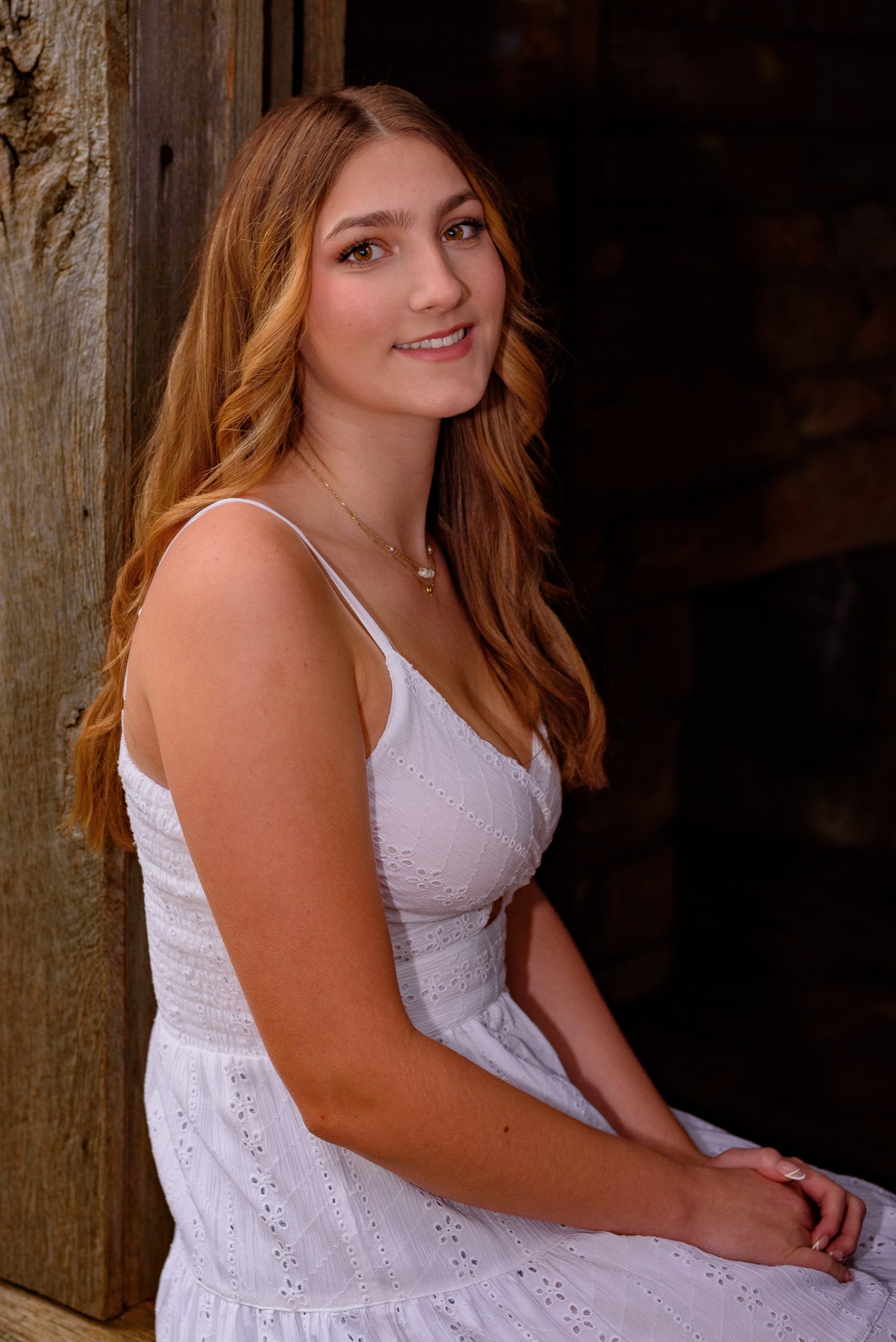 A young woman with long, wavy light brown hair wearing a white sleeveless dress sits indoors by a wooden wall, smiling softly at the camera—a look that embodies the fresh confidence of About Wake Skincare.