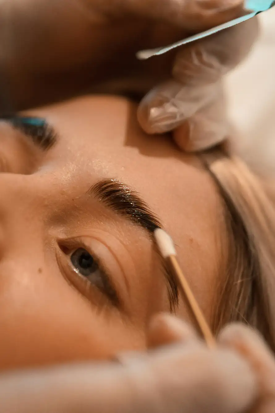 A close-up of a person's face as their eyebrows are being groomed. Gloved hands hold a cotton swab to the brow while another tool trims or shapes the eyebrow. The person’s eyes are open and looking upward.