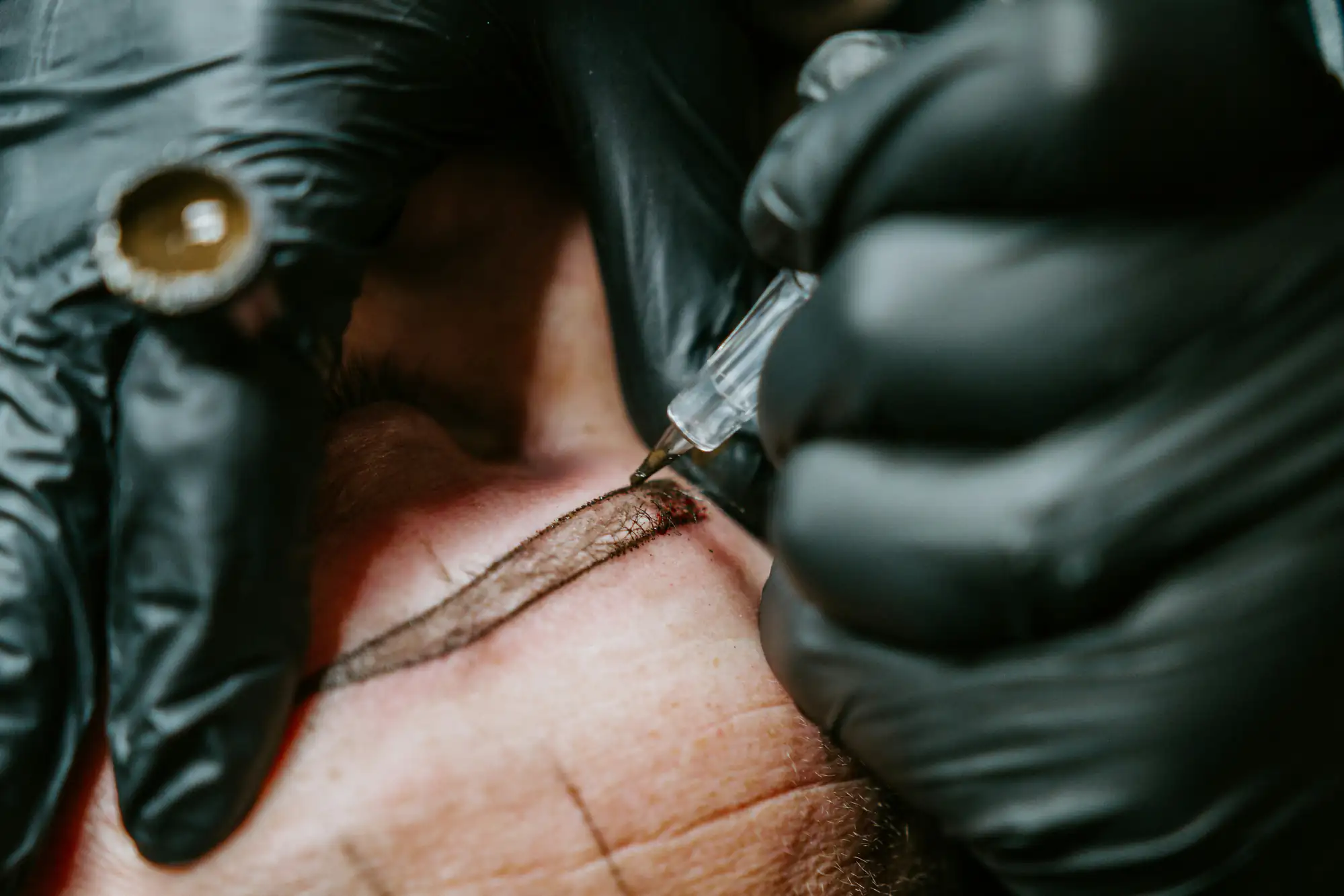 A close-up of a person receiving a cosmetic eyebrow tattoo, with a gloved hand holding a tattoo machine and carefully applying pigment along their eyebrow.