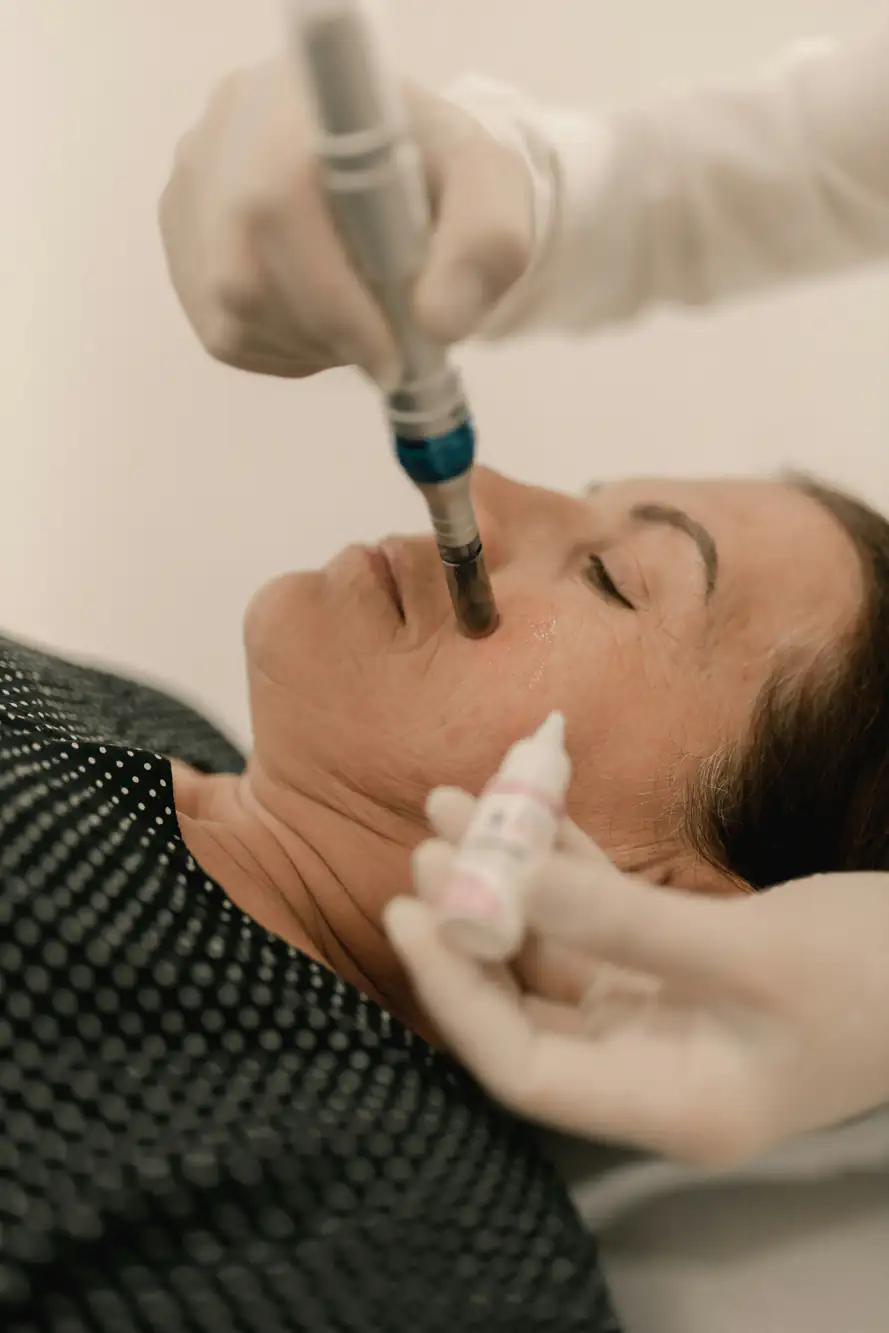 A woman lies on her back while a specialist wearing gloves performs a facial procedure using a handheld device on her cheek, and holds a small bottle of serum near her face.