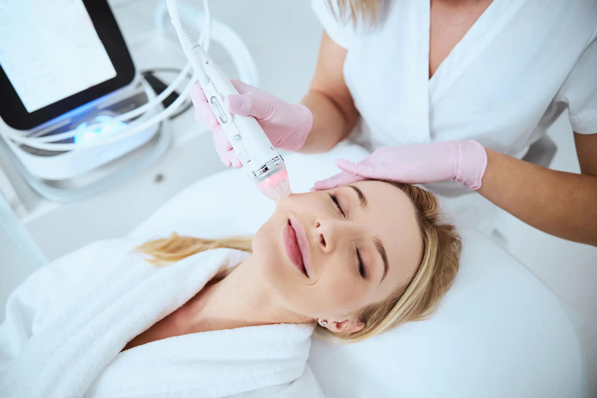 A woman in a white robe lies on a spa bed with her eyes closed and smiling as a skincare professional in gloves uses a facial device on her cheek during a beauty treatment.