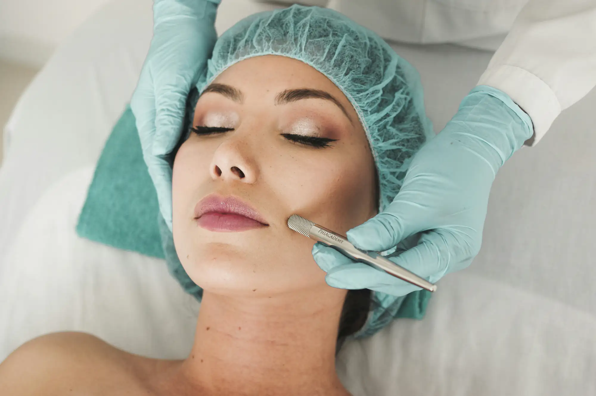 A woman lies on a treatment table with her eyes closed as a professional in gloves and a hair cap performs a facial procedure using a metal tool on her cheek.