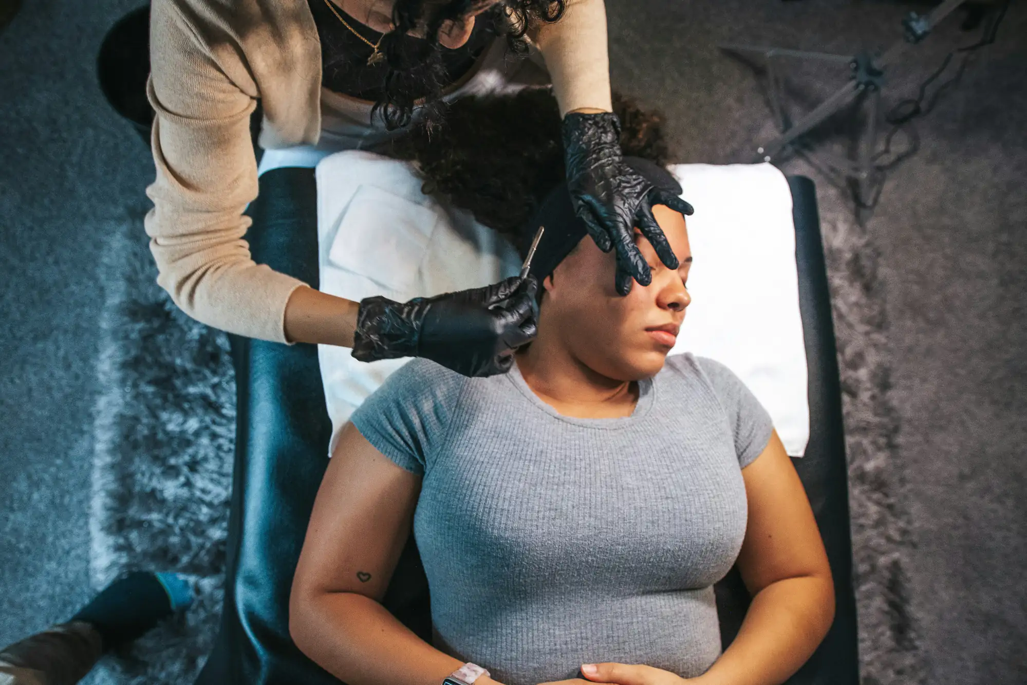 A woman lies on a padded table with her eyes closed while another person wearing black gloves shapes or grooms her eyebrows using tweezers. The setting appears to be a beauty or spa treatment room.